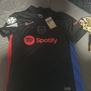 FC Barcelona bortatröja Lamine Yamal 19 - Säljer en svart FC Barcelona bortatröja från Nike med Lamine Yamal och nummer 19 på ryggen. Tröjan har röda och blå detaljer på sidorna, Champions League-märke på ärmen och Spotify-logga på bröstet.