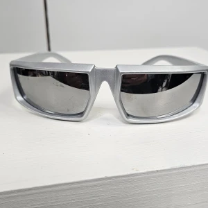 Silvriga futuristiska solglasögon - Snygga solglasögon med bred, silvrig båge och spegelglas. Modellen har en kantig, sportig design med markerad näsbrygga och breda skalmar. Perfekt för dig som vill sticka ut med en futuristisk look.