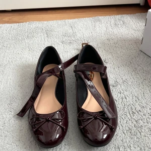 Vinröda lackade ballerinaskor med rosett - Säljer ett par vinröda ballerinaskor i lack med fina rosetter framtill och justerbara remmar över foten. Skorna har rund tå och platt sula, perfekta för en stilren look. Nypris 400:- mitt pris 200:-