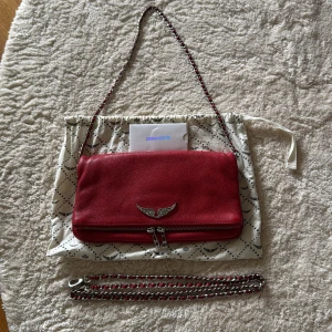 Röd väska från Zadig & Voltaire - Röd axelväska från Zadig & Voltaire (modell: Rock) med silverfärgade metalldetaljer. Medföljer det som visas på bild. Lite skavanker här och där - både på själva väskan, kanterna och remmarna. Finns även några mörkare ”fläckar” på baksidan. Köpt på Plick och har endast använt den ca 2 ggr. Endast seriösa köpare tack!!