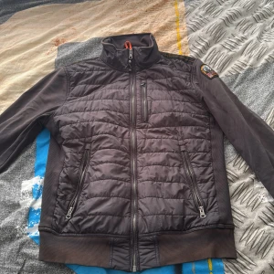 Svart quiltad jacka från Parajumpers - Snygg svart quiltad jacka från Parajumpers med hög krage och ribbade muddar. Jackan har dragkedja framtill och två snedställda fickor med dragkedja. Märkespatch på ena ärmen och sportig look.