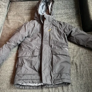 Grå vinterjacka med huva från Didriksons - Säljer en grå vinterjacka från Didriksons med huva och flera fickor. Jackan är 100% vattentät och har mysigt foder upptill på insidan. Perfekt för kalla dagar och håller dig torr i regn och snö.