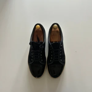 Svarta sneakers från Lanvin med lackad tå - Säljer ett par svarta sneakers från Lanvin med snygg lackad tå och ovandel i mocka. Skorna har snörning och vit platt sula. Perfekta för dig som gillar stilrena och exklusiva sneakers med en twist.