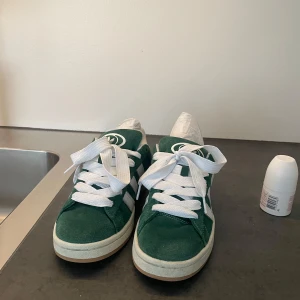 Gröna Adidas Campus sneakers - Säljer ett par gröna Adidas Campus sneakers med vita ränder och vit sula. Skorna har klassisk snörning och är tillverkade i mocka med detaljer i vitt läder. Perfekta för dig som gillar retrostil och streetwear. Pris kan diskuteras 