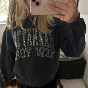 Mörkgrå sweatshirt med strasstext - Säljer en mörkgrå sweatshirt med texten 'Manhattan New York' i glittriga strasstenar på bröstet. Tröjan har rund hals och långa ärmar, perfekt för en avslappnad och cool stil.