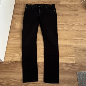 Svarta jeans från Nudie Jeans - Säljer ett par svarta jeans från Nudie Jeans med klassisk femficksdesign och raka ben. Jeansen har dragkedja och knapp i midjan samt diskreta sömmar. Perfekta för dig som gillar stilrena och tidlösa byxor.