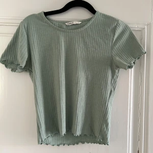 Ljusgrön ribbad topp från ONLY - Säljer en ljusgrön ribbad topp från ONLY med korta ärmar och vågiga kanter vid ärmslut och nederkant. Toppen har en rund halsringning och är i mjukt, stretchigt material.