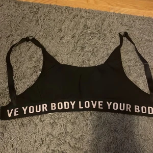 Svart BH från Esotiq - Svart linnetopp från Esotiq med bred resår nertill där det står 'LOVE YOUR BODY' i stora bokstäver. Justerbara axelband och hakknäppning bak. Perfekt för träning eller chill, i mjukt och stretchigt material. Det är storlek S, 