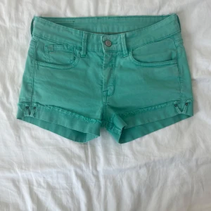 Turkosa lågmidjade jeansshorts  - Snygga turkosa lågmidjade shorts från Le Temps des Cerises med råa kanter och klassisk femficksmodell. Shorts med låg midja, bälteshällor och dekorativa nitar framtill. Perfekta för varma sommardagar och chill häng. 