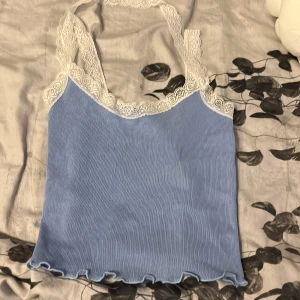 Ljusblå ribbad linnetopp med spets - Supersöt ljusblå linnetopp med ribbad struktur och vita spetsdetaljer längs urringningen och axelbanden. Toppen är ärmlös och har en lätt vågig kant nertill som ger en extra touch. Perfekt för dig som gillar en romantisk och trendig stil.