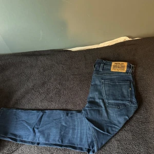 Blå Replay Anbass jeans - Snygga blå jeans från Replay, modell Anbass. Klassisk femficksdesign med tydlig Replay-patch bak i midjan. Jeansen har en mörk tvätt och tillverkade i mjukt denimtyg som sitter skönt hela dagen. Enda defekten syns på bild 4.