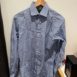 Blå randig skjorta Polo Ralph Lauren - Klassisk blå skjorta från Polo Ralph Lauren med vita och ljusblå ränder. Skjortan har lång ärm, knappar framtill och en snygg krage. Perfekt för en stilren och avslappnad look.