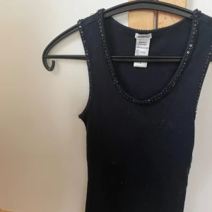 Svart linnetopp med paljetter från Guess - Snygg svart linnetopp från Guess med ribbad struktur och glittriga paljettdetaljer längs halsringning och ärmhål. Tillverkad i mjuk bomull med polyesterkant, perfekt för att lyfta din outfit med lite extra bling.