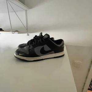 Nike Dunk sneakers i svart och grå - Nike Dunk SB sneakers i svart och grå med neongula detaljer och logga. Skorna har lågt skaft, snörning med svarta och vita skosnören samt en vit sula. Ovandelen är i mocka och syntet, med klassisk Nike Swoosh på sidan och broderad logga bak.