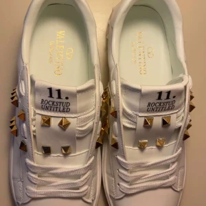 Valentino Rockstud vita sneakers - Snygga vita Valentino Rockstud sneakers med guldiga nitar och coola detaljer. Skorna har rund tå, platt sula och är gjorda i skinn. Klassisk snörning och ikoniska pyramidnitar längs sidorna och bak på hälen. Perfekta för dig som vill sticka ut med lyxig streetstyle.