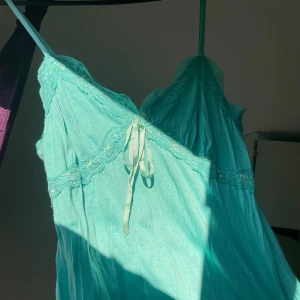 Turkos topp med spets och rosett - Supersöt turkos topp med justerbara tunna axelband, spetsdetaljer längs byst och nederkant samt en liten rosett framtill. Toppens tyg är lätt och luftigt, perfekt för varma dagar. Snygg v-ringning och romantisk känsla.