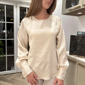 Beige blus från Vero Moda med knappar - Snygg beige blus från Vero Moda i lätt skimrande material. Blusen har långa puffärmar med breda manschetter och tre klädda knappar. Rund halsringning och knappslå i nacken. Perfekt för dig som gillar stilrena och trendiga plagg.