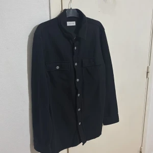 Svart overshirt från Pier One - Stilren svart overshirt från Pier One med klassisk krage och raka linjer. Skjortan har två stora bröstfickor med lock och knappar framtill. Tillverkad i ett mjukt material som känns lite ulligt, perfekt för lager-på-lager. Långärmad och med snygga detaljer på fickor och ärmslut. Säljer pågrund av att den är för stor, priset går o diskutera