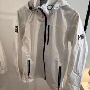Vit vindjacka från Helly Hansen - Snygg vit vindjacka från Helly Hansen med svart dragkedja och röda detaljer. Jackan har huva, meshfoder och flera fickor med dragkedja. Perfekt för blåsiga dagar och har en clean look med HH-logga på ärmen.