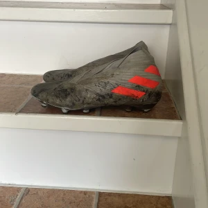 Adidas Nemeziz grå fotbollsskor - Säljer ett par gråa Adidas Nemeziz fotbollsskor med orangea detaljer och klassiska tre ränder på sidan. Skorna har en strömlinjeformad design utan snörning och är tillverkade i syntetmaterial. Perfekta för dig som vill sticka ut på planen.