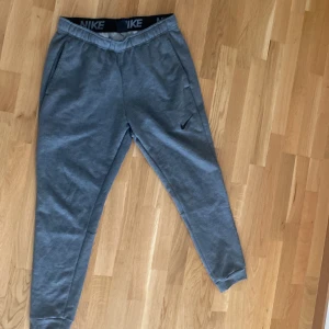 Grå Nike Dri-FIT mjukisbyxor M - Grå mjukisbyxor från Nike med Dri-FIT-teknologi. Byxorna har resår i midjan, snedställda fickor och en diskret Nike-logga på benet. Perfekta för chill eller träning, med en skön och sportig passform.