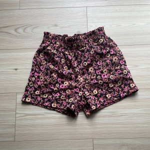 Blommiga shorts från Lindex - Snygga shorts från Lindex med blommigt mönster i rosa, beige och svart. De har elastisk midja för extra komfort och är perfekta för varma dagar. Mjukt bomullsmaterial som känns skönt mot huden och ger en avslappnad vibe. I storlek 134-140.