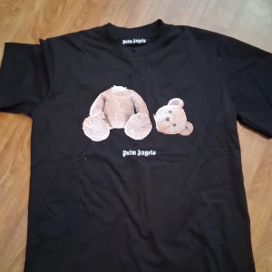 Palm Angels svart t-shirt med björntryck - Svart t-shirt från Palm Angels med ikoniskt björntryck framtill och stor logga på ryggen. Klassisk passform och rund halsringning. Materialet är mjuk bomull som känns skönt mot huden. Perfekt statement-plagg för dig som gillar streetwear och coola prints.