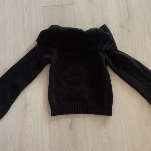Svart offshoulder stickad tröja - Svart stickad tröja med bred, ribbad offshoulder-krage. Långärmad modell med ribbade muddar vid ärmslut och nederkant. Perfekt för dig som vill ha en stilren och mysig look med lite edge.