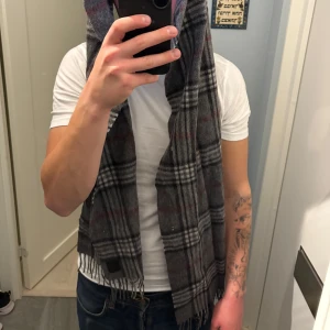 Rutig grå halsduk från Amanda Christensen - Snygg och klassisk rutig halsduk från Amanda Christensen i grått med inslag av svart och röda linjer. Halsduken är mjuk och har fransar i ändarna.