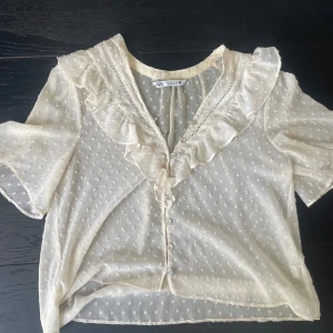 Vit blus med volang från Zara - Supersöt vit blus från Zara med korta ärmar, volang längs v-ringningen och små prickar i tyget. Blusen är genomskinlig och har knappar. Perfekt för dig som gillar en feminin och trendig stil. Frakt ingår ej 
