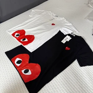 Vit Comme des Garçons Play t-shirt hjärta - Säljer en Comme des Garçons Play t-shirt i vitt med det ikoniska röda hjärta-trycket med ögon på magen och ett mindre broderat hjärta på bröstet. Klassisk passform, rund hals och korta ärmar. Materialet är mjuk bomull, perfekt för en clean och cool look.