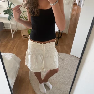 Beige kortkjol med snörning och volang - Supersöt beige kortkjol med smock i midjan och snörning med tofsar. Kjolen har volangkant upptill och ett diskret mönster i tyget. Några få stenar på snöret har lossnat men det syns inte tydligt, se bild 4🤍