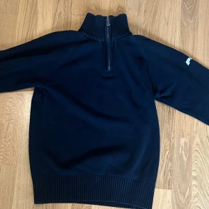 Mörkblå stickad Peak Performance-tröja - Mörkblå stickad tröja från Peak Performance med hög ribbad krage och half zip-dragkedja. Broderad logga på ärmen. Perfekt för kyliga dagar och snygg till jeans eller cargobyxor. 
