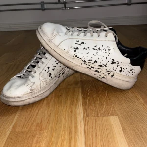 Vita sneakers med svart stänkmönster - Snygga vita sneakers med svart stänkmönster och svarta detaljer vid hälen. Skorna har klassisk snörning och platt sula, perfekt för en streetstyle-look. Materialet är troligtvis skinn eller syntet. Unik design som sticker ut. Det är skor från Zara