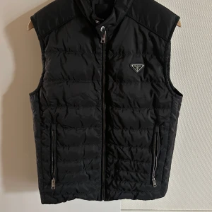 Svart dunväst från Prada - Snygg svart dunväst från Prada med quiltad design och klassisk logga på bröstet. Västen har hög krage, två fickor med dragkedja och stängs med dragkedja framtill. Perfekt för lager-på-lager och stilren look. Har använt kanske 1-2 gånger. Pris kan diskuteras 