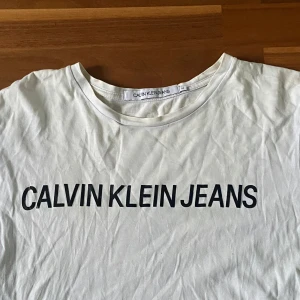 Vit Calvin Klein Jeans t-shirt - Vit t-shirt från Calvin Klein Jeans med svart logotyptryck över bröstet. Klassisk rund halsringning och korta ärmar. Tillverkad i mjuk bomull, perfekt för en clean och stilren look.