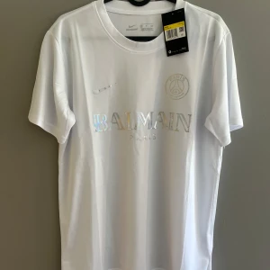 Vit Balmain x PSG t-shirt Nike - Vit t-shirt från Balmain x PSG i samarbete med Nike. T-shirten har korta ärmar, rund hals och holografiskt tryck med Balmain Paris och PSG-logga på bröstet. Tillverkad i ett lätt och svalt polyestermaterial, perfekt för en clean och sportig look.