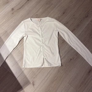 Vit ribbad topp med rosetter från Nelly - Supersöt vit ribbad topp från Nelly i storlek XS. Långärmad med flera små rosetter framtill. Tröjan är i nyskick. Köparen står för frakten