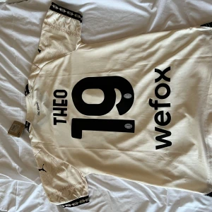 AC Milan bortatröja Puma  - AC Milan bortatröja från Puma med Theo och nummer 19 på ryggen. Tröjan är ljusbeige med svarta detaljer, korta ärmar och subtilt mönster inspirerat av gotisk arkitektur. Broderat klubbmärke på bröstet och svarta muddar med stjärnmönster. Tillverkad i lätt och ventilerande material. Storlek M