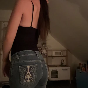 Blå bootcut jeans med broderad ficka - Snygga blå bootcut jeans med broderad bakficka i beige och lila mönster. Jeansen har klassisk femficksdesign, dragkedja och knapp framtill. Perfekta för dig som vill ha en unik detalj på dina jeans och gillar en lite utsvängd passform. Står inte storlek eller var dom är ifrån men är rätt säker på att dom är xs. 