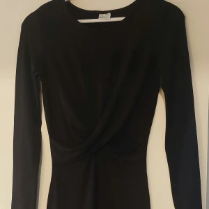 Svart långärmad klänning från Gina Tricot - Snygg svart kortklänning från Gina Tricot med lång ärm och twistad detalj framtill i midjan. Klänningen har en figurnära passform och är tillverkad i ett mjukt, stretchigt material som sitter bekvämt på kroppen.