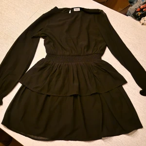 Svart volangklänning från Kappahl - Supersnygg svart kortklänning från Kappahl med långa ärmar och markerad midja med smock. Klänningen har dubbla volanger i kjolen som ger en extra flowig känsla. Perfekt för dig som gillar en stilren look med lite extra detalj.