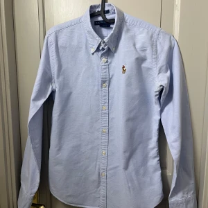 Ljusblå skjorta från Ralph Lauren - Klassisk ljusblå skjorta från Ralph Lauren i slim fit-modell. Skjortan har knappar framtill och den ikoniska broderade loggan på bröstet. Tillverkad i mjuk bomull och passar perfekt till en stilren och avslappnad look. Tveka inte på att höra av er vid frågor eller funderingar.