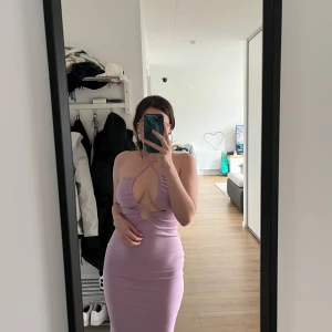 Ljuslila långklänning från Gina Tricot - Supersnygg långklänning i ljuslila från Gina Tricot. Klänningen har en tight passform, är ärmlös och har en öppen rygg med snygga knytband i nacken. Framtill finns en cut-out detalj med ring, vilket ger en trendig look. Perfekt för dig som vill sticka ut.