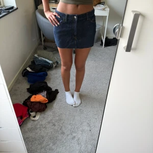 Mörkblå jeanskjol med rå kant - Snygg mörkblå jeanskjol i kort modell med klassiska fickor fram och bak. Kjolen har en rå, avklippt kant nertill och stängs med knapp och dragkedja framtill. Perfekt för dig som gillar en enkel och trendig denimlook.