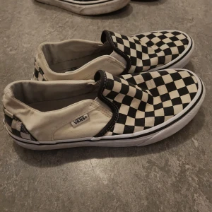 Vans slip-on checkerboard sneakers - Knappt använda, tvättad. Klassiska Vans slip-on sneakers med svartvitt rutigt mönster. Skorna har låg profil, platt sula och är tillverkade i canvas. Perfekta för en avslappnad streetstyle-look och superenkla att bara hoppa i.