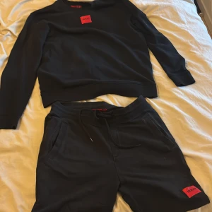 Svarta shorts och tröja från Hugo med röd logga - Svarta shorts från Hugo med en tydlig röd logga på vänster ben. Mjukt material, elastisk midja med snörning och två sidofickor. Perfekta för chill dagar eller när du vill ha en clean och stilren look. Kläderna kan säljas separat 