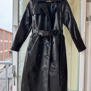 Day Birger et Mikkelsen DK - Trenchcoat - Säljer en svart, glansig trenchcoat i skinnlook med bälte i midjan och dubbelknäppning. Jackan har långa ärmar, klassisk krage och är tillverkad i polyester och polyuretan. Perfekt för dig som vill sticka ut med en edgy vibe. Det saknas 1 knapp på framsidan men går lätt att knäppa tät med de andra knapparna och bältet åtdraget. Inhandlad på NK 2019 för ca 4000kr. 