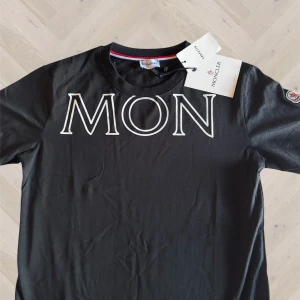 Svart Moncler t-shirt med logga - Svart t-shirt från Moncler med stor vit logotyp över bröstet. Klassisk rund halsringning med röd och vit detalj på insidan av kragen. Kortärmad modell i mjuk bomull, perfekt för en clean och stilren look.
