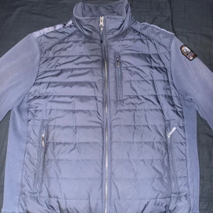 Blå pufferjacka från Parajumpers - Snygg blå pufferjacka från Parajumpers med quiltad framsida och rygg, samt mjuka ärmar. Jackan har hög krage, flera dragkedjefickor och en patch på ärmen. Perfekt för dig som vill ha en stilren och sportig look. Har ett yttepytte litet hål på vänstra sida ficka men sysn det inte på bilderna syns det inte heller i verkligenheten!💯